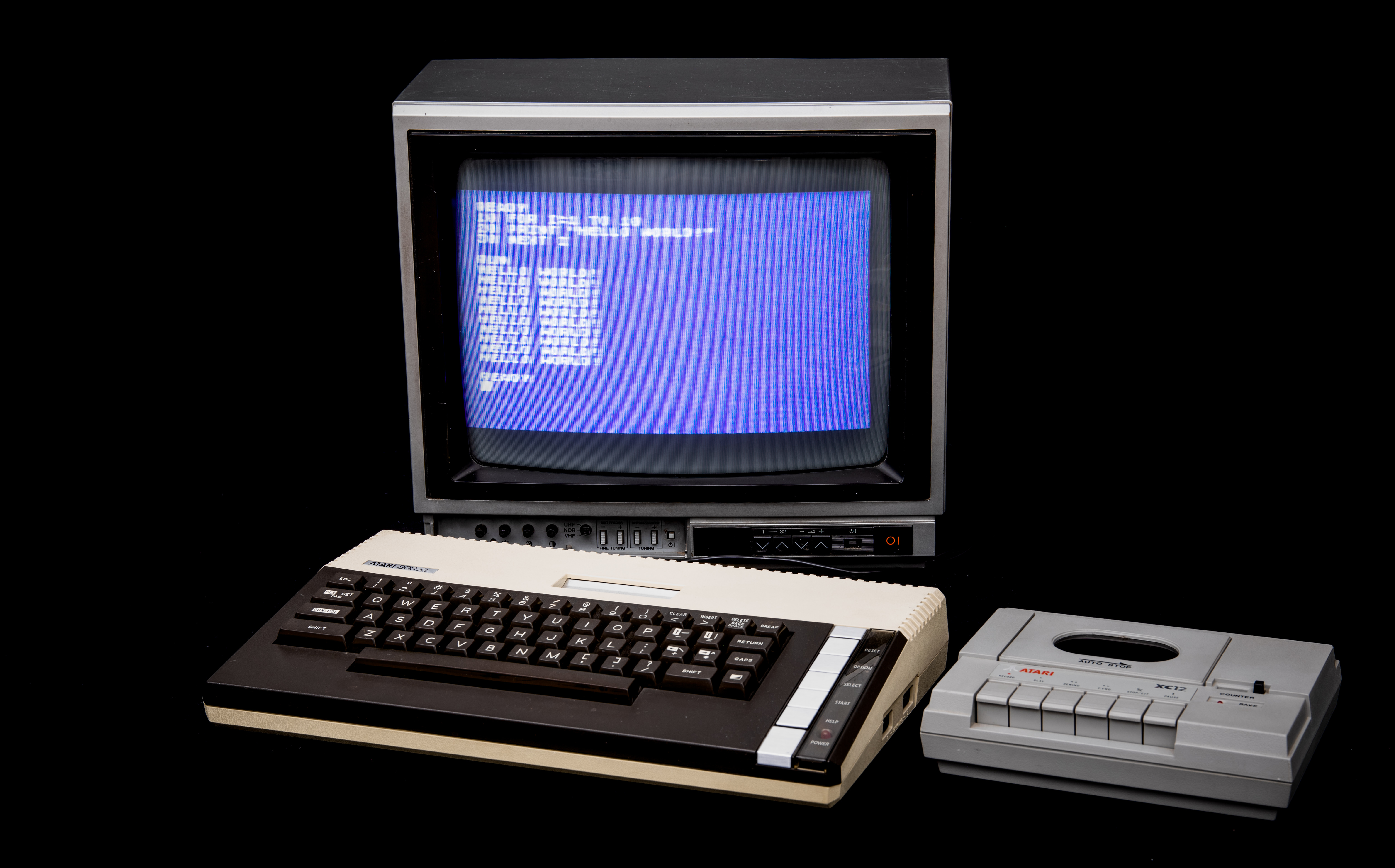 An Atari 800XL computer and XC12 cassette recorder, with a CRT monitor showing a running BASIC program that prints HELLO WORLD! in a loop